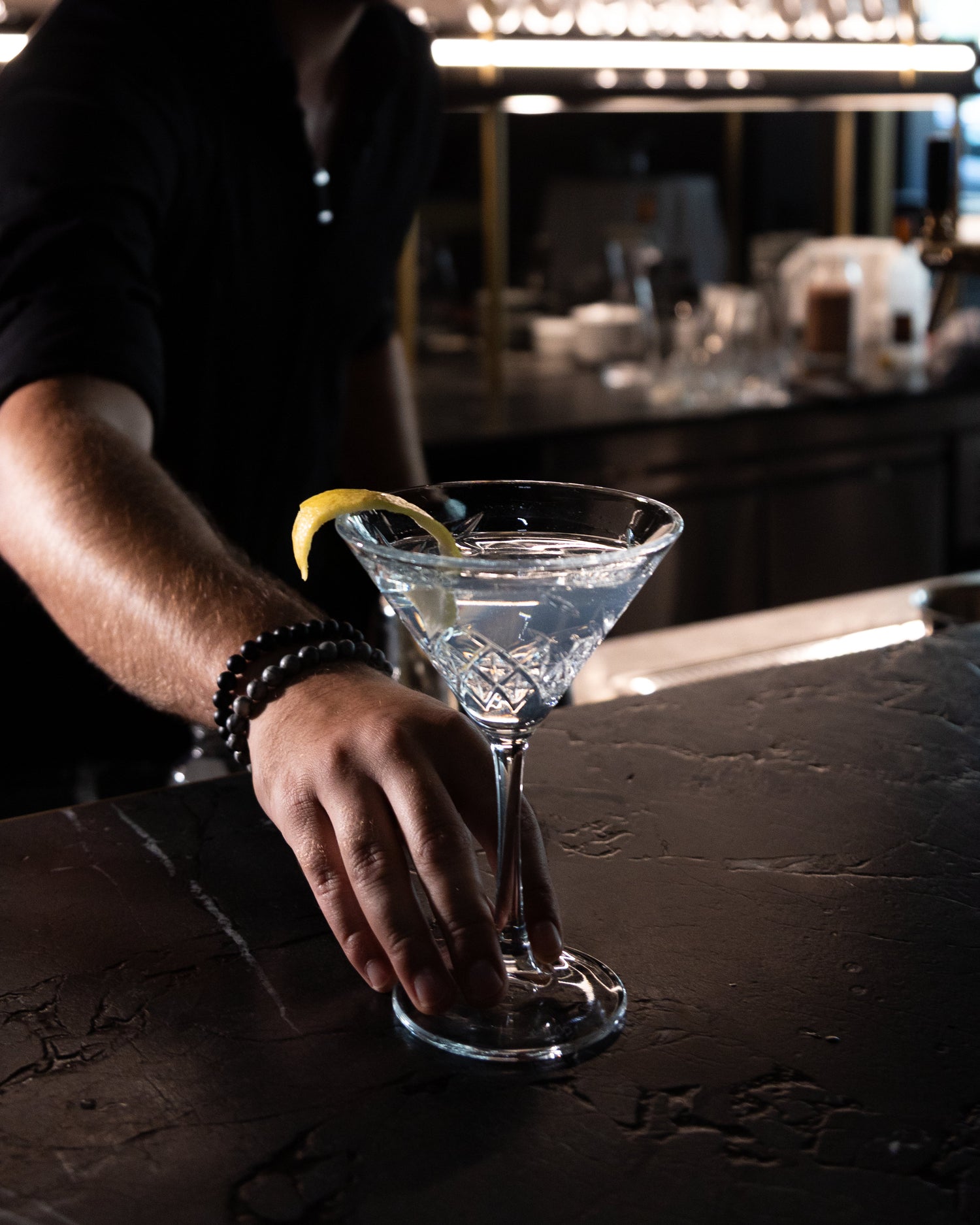 Gin präsentiert in einem Martini Glas uns einer Zitronenscheibe. Das Glas wird von einer Hand über einen Tresen geschoben